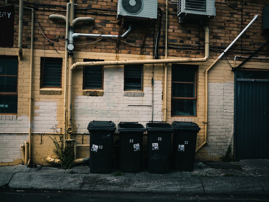 A large pile of black garbage bags, some torn open revealing visible waste, is stacked in front of a red metal door set into a concrete wall. The door features various graffiti tags in white and pink 