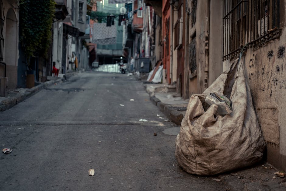 A street scene showing a white waste collection truck with an open rear hopper, positioned alongside a sidewalk in front of multi-story residential buildings with aged facades and small windows. A wor